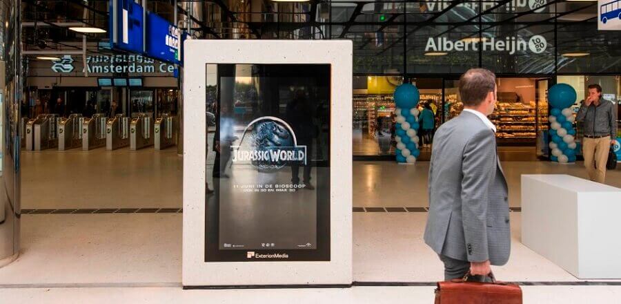 A man walking by an Exterion screen in Amsterdam Central Station. The screen is playing an ad for Jurassic World.
