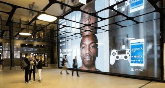A giant digital screen in a train station lobby. On the screen is an ad for Sony PlayStation and Sony phones.