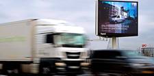 A digital billboard off to the side of a highway. It advertises a corporate minivan sold by Peugeot.