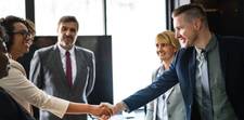A group of people around a meeting table. A man and a woman are shaking hands, implying a deal has been made.