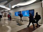 Travelers walking through an airport concourse past large digital out-of-home advertising screens displaying weather information and airport messaging.