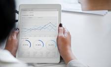 A woman holding a tablet. On the tablet's screen are charts and figures relating to finances. Reviewing data in this way is an important method of determining DOOH campaign effectiveness.