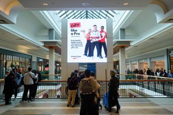 A busy two-level shopping mall atrium with shoppers gathered along the upper-floor railing. A large digital billboard dominates the center, displaying a SportChek holiday advertisement beneath a glass skylight.