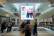 A busy two-level shopping mall atrium with shoppers gathered along the upper-floor railing. A large digital billboard dominates the center, displaying a SportChek holiday advertisement beneath a glass skylight.