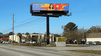 A roadside scene featuring a large Chumba Casino billboard above a Dollar General store. The billboard has a bright orange-and-yellow design with a smiling woman holding a phone and the text, “Hot Dang! As Tasty as Tennessee Chicken,” against a clear blue sky.