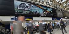 Transit users walking through Waterloo station. Overhead, a large digital billboard displays an ad for the film &quot;Jurassic World.&quot; Transit hubs are high-traffic areas perfect for connecting people with favourite brands.