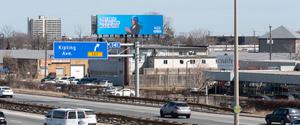 <p>A busy roadway. Above and in the background, a Pattison digital billboard displays an ad.</p>
