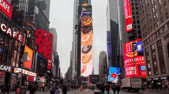 A bustling Times Square streetscape featuring a towering digital Crumbl advertisement displaying colorful cookie varieties, surrounded by illuminated billboards, skyscrapers, pedestrians, and city traffic.