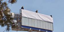 Three workers on top of a billboard, about to unfurl a poster that is due to be posted