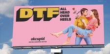 A bright pink billboard showing two women embracing, one holding a long-stemmed red rose in her hand. The heading text says &quot;DTFall Head Over Heels.&quot;