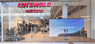 Exterior view of a Cotswold Outdoor store in a shopping centre, featuring a large digital display in the storefront showing an outdoor campaign.